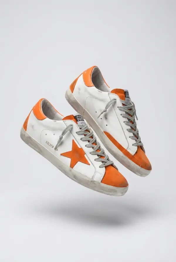 A pair of Golden Goose Superstar sneakers floating in mid-air against a plain white background. The shoes feature white leather uppers with the brand's signature distressed and scuffed finish.